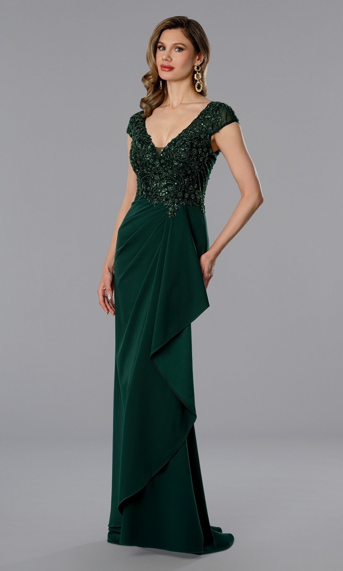 A woman models the Long Formal Dress: Stella Couture 25364 in Hunter, featuring a beaded v-neck bodice, cap sleeves, and a draped skirt, standing against a plain gray background.